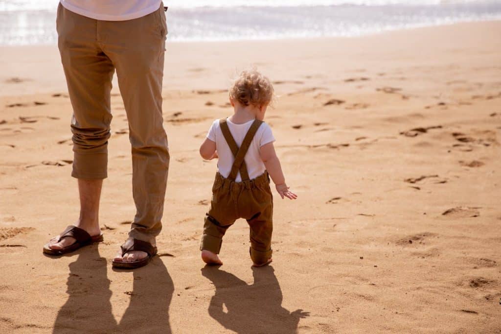 Toddler walking on sandy beach with faceless father